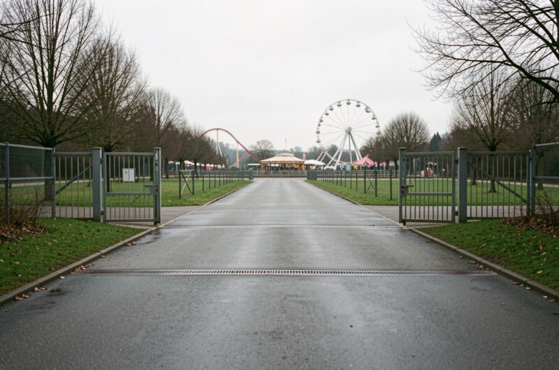 Vue réaliste de l’entrée calme d’un parc de loisirs fermé en Moselle, avec des manèges visibles à l’arrière-plan sous un ciel couvert.
